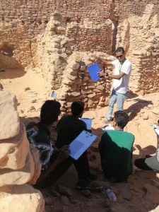 Teacher or guide explains ancient stone ruins to students with clipboards in a desert site outdoors.