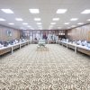 A large conference room with people seated around U-shaped tables, flags lining the back wall, and a patterned carpet under bright ceiling lights.
