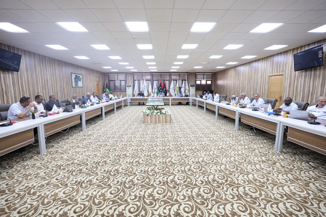 A large conference room with people seated around U-shaped tables, flags lining the back wall, and a patterned carpet under bright ceiling lights.