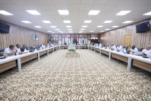 A large conference room with people seated around U-shaped tables, flags lining the back wall, and a patterned carpet under bright ceiling lights.