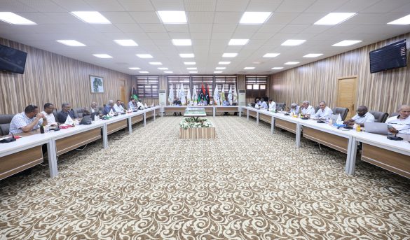 03EBB295-92B1-49B3-9FEC-A9A2466EC2A3 - جامعة سبها | Sebha University A large conference room with people seated around U-shaped tables, flags lining the back wall, and a patterned carpet under bright ceiling lights.