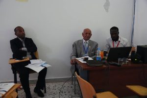 05BB1372-92A8-4E72-92C0-B808B5C7B8CC - جامعة سبها | Sebha University Three men sit in a small meeting room; a senior man in a gray suit is at the desk with a laptop, flanked by two coworkers.