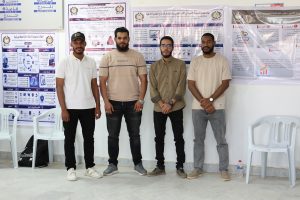 Four men stand side by side indoors in front of large Arabic posters on a wall, posing for a photo near plastic chairs.
