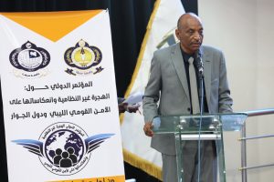 Man in a gray suit speaks at a glass podium during a conference, with a large banner and logos behind him.