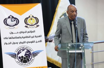 Man in a gray suit speaks at a glass podium during a conference, with a large banner and logos behind him.