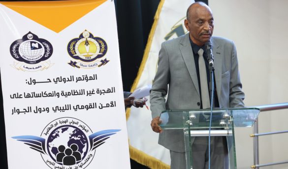 0862814A-09BD-44E1-B712-CBA1BC47B345 - جامعة سبها | Sebha University Man in a gray suit speaks at a glass podium during a conference, with a large banner and logos behind him.