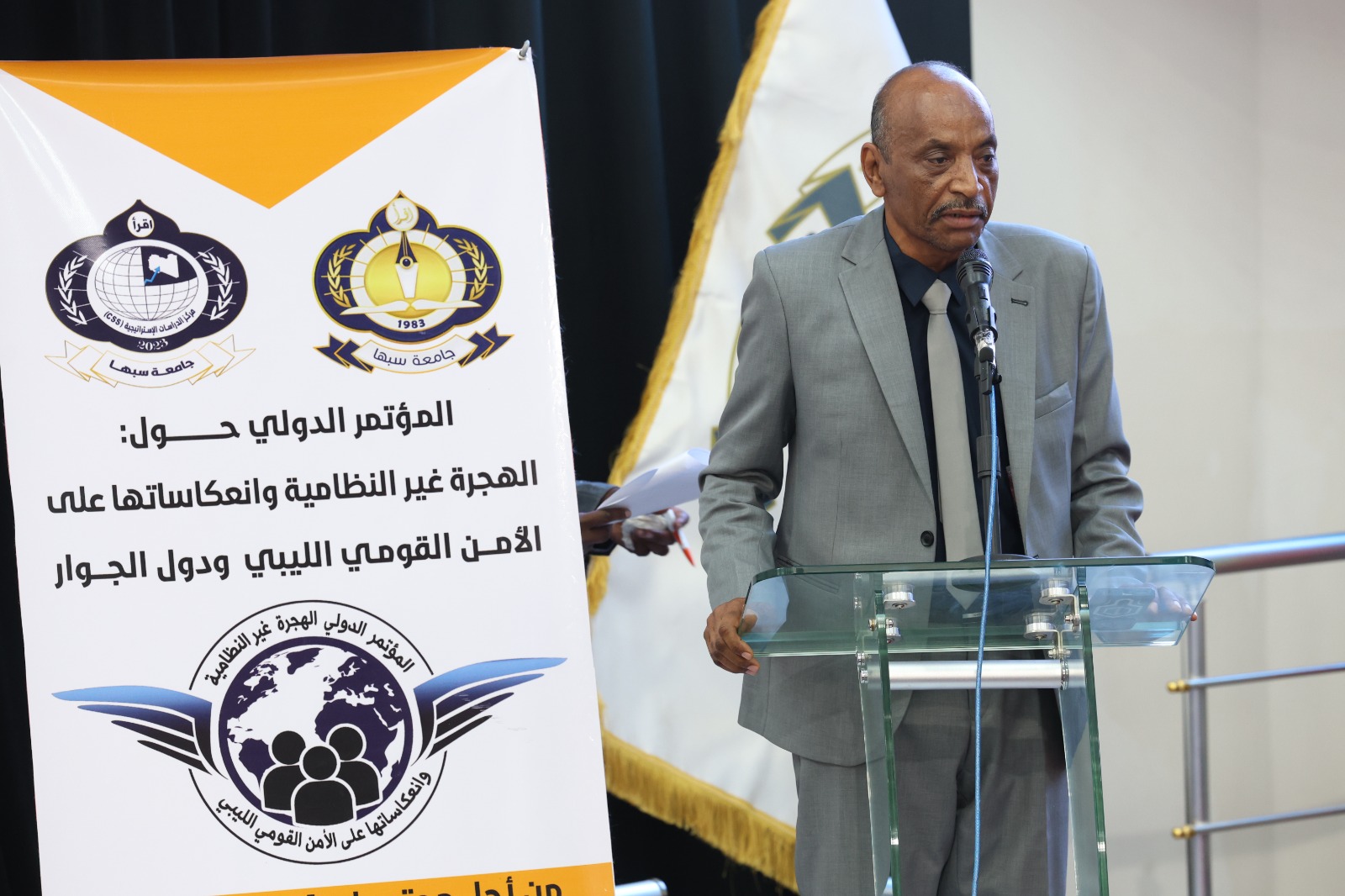 Man in a gray suit speaks at a glass podium during a conference, with a large banner and logos behind him.