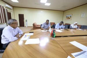 0956ED4A-7EE6-4DA9-B957-D323365F0638 - جامعة سبها | Sebha University Four adults sit around a large wooden conference table, reviewing papers; small flags and scattered documents on the table in a beige-walled room with framed art in the background.
