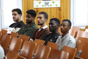 Group of young men seated in a classroom, looking toward the front, wood-paneled wall behind them.