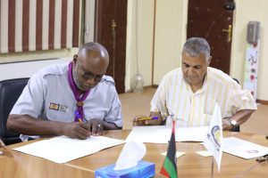 Two men sign documents at a table during a formal meeting, with flags and tissues in the foreground.