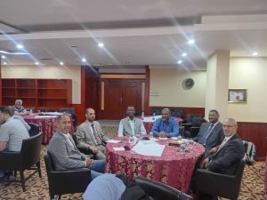 0BFB9F5E-434F-4D4B-B1D3-2C6DA39AFA57 - جامعة سبها | Sebha University Group of men in suits seated around a round table in a conference room, discussing with papers and bottles on the table.