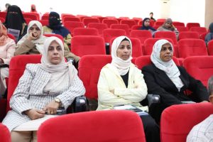 0C0E9EEC-91C0-470F-B2C6-97541502EE7E - جامعة سبها | Sebha University Three women wearing hijabs seated in a red theater-style auditorium watching an event in progress.