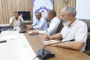 0C23D363-D0DC-4646-8E36-C0BF43F5FBEF - جامعة سبها | Sebha University Four men sit around a conference table; one on the left raises his hands while speaking, others listen.