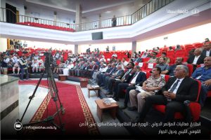 Audience seated in red theater chairs watching a formal conference in a large auditorium; camera operator and equipment in foreground, UAE-style stage area visible.