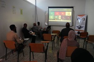 0E34F3F8-E296-4DBD-B93B-DB7EA3A3ED51 - جامعة سبها | Sebha University Panel of professionals in a classroom watching a video conference on a projector screen showing a grid of participants; a banner with logos stands to the right.