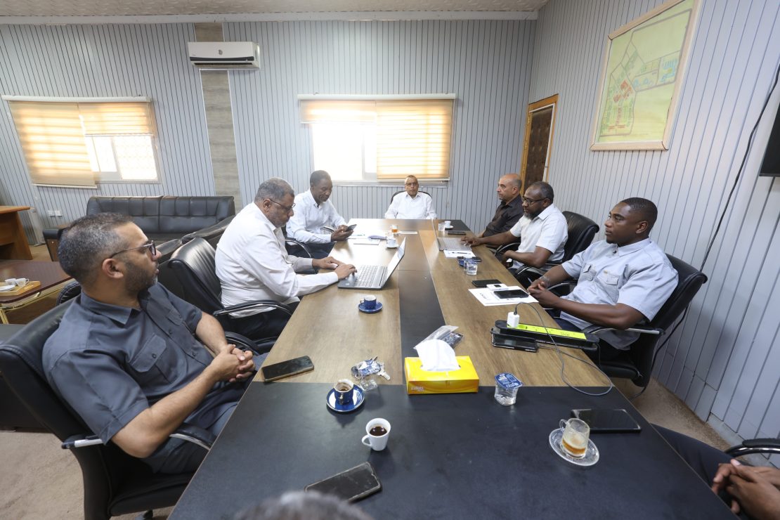 Group of men in business attire seated around a long conference table in a meeting room, with laptops and coffee cups.
