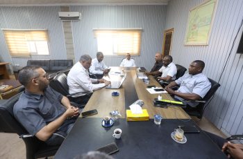 Group of men in business attire seated around a long conference table in a meeting room, with laptops and coffee cups.