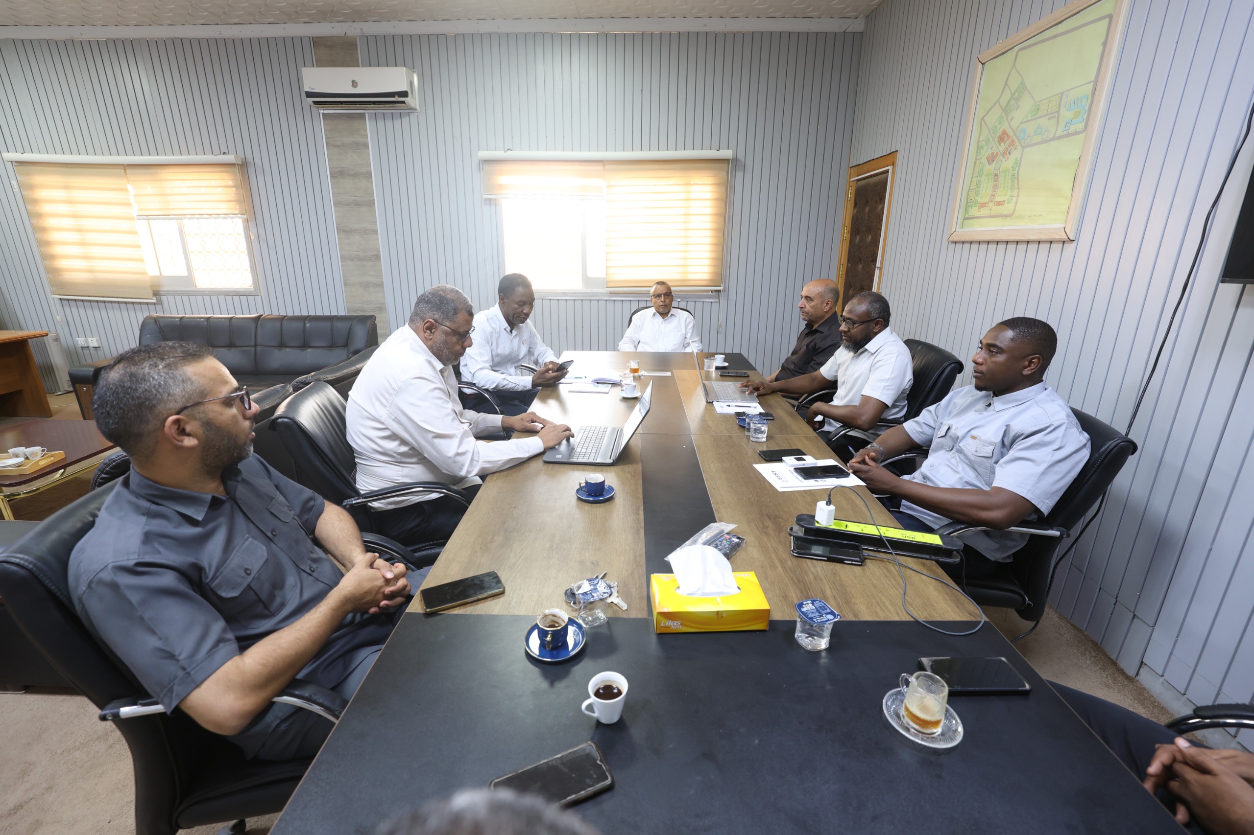 Group of men in business attire seated around a long conference table in a meeting room, with laptops and coffee cups.