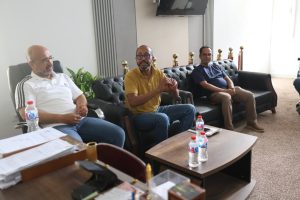 1293EC1F-73B8-4D93-8A2C-E2BE2632AB49 - جامعة سبها | Sebha University Three men sit on black leather couches in a modern office, chatting around a wooden coffee table with bottled water and papers nearby.