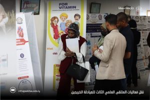 Group of people at a poster exhibition or science fair, with posters about vitamins; a woman in a hijab reads a document in the foreground.
