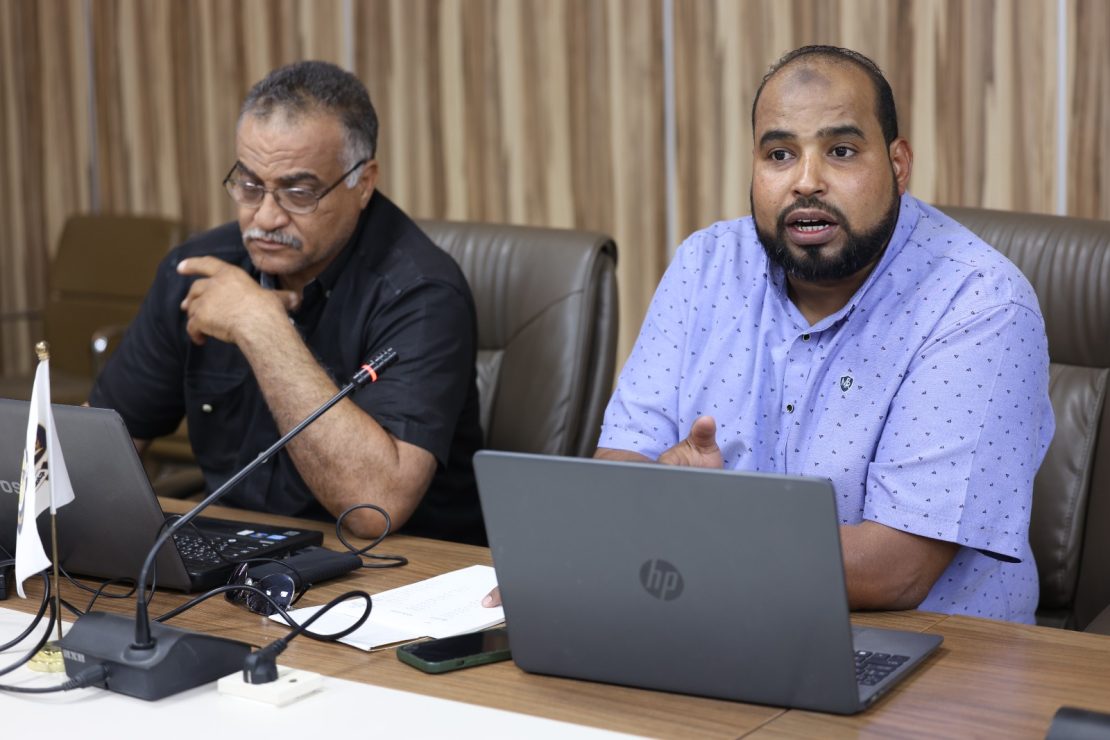 Two men seated at a conference table with laptops, one speaking into a microphone as the other looks on.