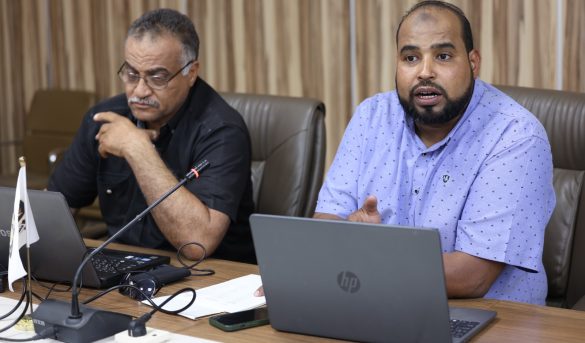Two men seated at a conference table with laptops, one speaking into a microphone as the other looks on.