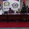 Panel of five people seated at a conference table in a Sabha University event with banners behind them and Libyan flags nearby, serious discussion in progress.