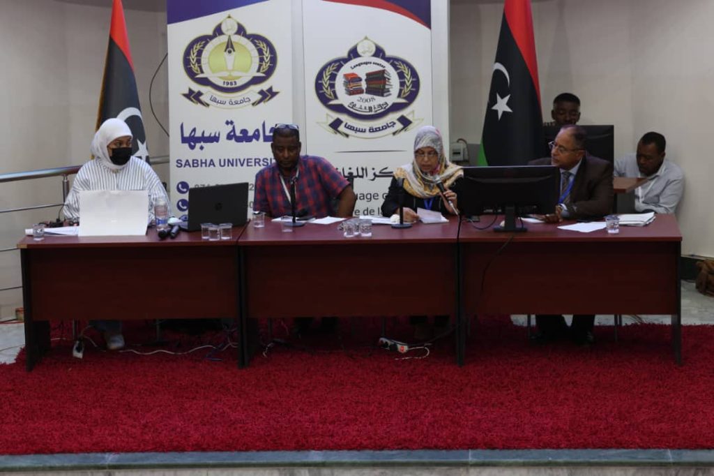 Panel of five people seated at a conference table in a Sabha University event with banners behind them and Libyan flags nearby, serious discussion in progress.