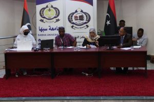 180A29D2-9297-49CB-B2C7-5CBC3DAA33FF - جامعة سبها | Sebha University Panel of five people seated at a conference table in a Sabha University event with banners behind them and Libyan flags nearby, serious discussion in progress.
