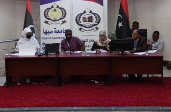 Panel of five people seated at a conference table in a Sabha University event with banners behind them and Libyan flags nearby, serious discussion in progress.