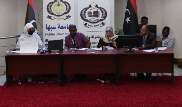 Panel of five people seated at a conference table in a Sabha University event with banners behind them and Libyan flags nearby, serious discussion in progress.