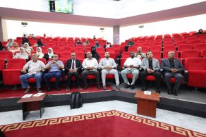 192CB832-713A-4461-9529-7CEAB81FD69F - جامعة سبها | Sebha University Group of formally dressed people seated in a red-carpeted conference hall, facing the stage in a dull-lit auditorium.