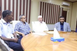 19BF9048-1E63-47FE-B64C-2BCCC5D4E1EF - جامعة سبها | Sebha University Four men seated around a conference table in a meeting room, with papers and a tissue box on the table, appears to be a formal discussion.