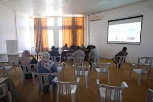 1A6A49E2-22D1-4801-B39D-CC4CF3A53239 - جامعة سبها | Sebha University Indoor training or meeting room with a panel at the front and attendees seated on white plastic chairs facing a projector screen with a slide; large windows with orange curtains to the left.