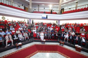 20E76823-73D6-4C7B-9B2A-354F2DD06125 - جامعة سبها | Sebha University Large conference hall full of people seated in red chairs, facing a stage with a patterned red carpet.