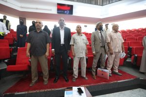 Group of men and women standing in a red-upholstered auditorium, some in suits, others in casual attire, facing forward; Turkish flag image on screen above.
