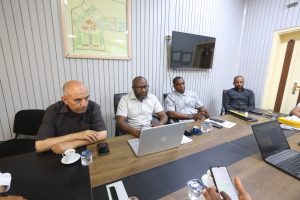 2538F400-DF73-4EBA-8FE5-E4EDA3D9163D - جامعة سبها | Sebha University Group of five men sit around a conference table with laptops, phones, and coffee cups in a bright office room.