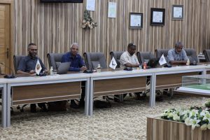 Five men sit at a long conference table in a formal meeting room with framed certificates on a wood-paneled wall.