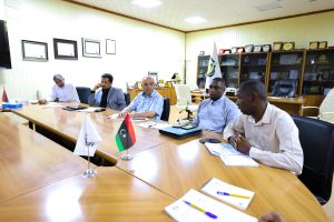 26E1D8D0-3328-41DD-B100-4F08A5516694 - جامعة سبها | Sebha University Group of men seated around a long conference table in a formal meeting room, with flags and awards on display in the background.