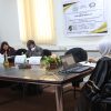 Group of people in a meeting around a long table in a conference room, with a poster on the wall and laptops in front of them.