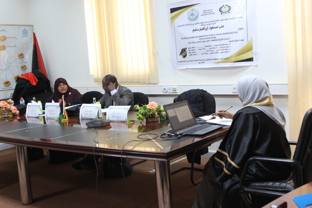 Group of people in a meeting around a long table in a conference room, with a poster on the wall and laptops in front of them.