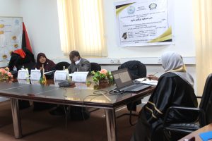 Group of people in a meeting around a long table in a conference room, with a poster on the wall and laptops in front of them.