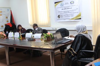 Group of people in a meeting around a long table in a conference room, with a poster on the wall and laptops in front of them.