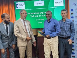 Four men in suits stand on a stage smiling as a certificate is handed over, with a green slide behind reading 'Pedagogical Engineering Training of Trainers'.