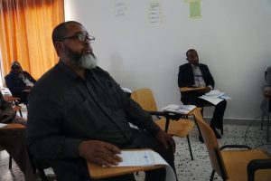 2B21A3FF-E23E-42B5-9610-40D865FAABB0 - جامعة سبها | Sebha University Bearded man with glasses in a dark shirt sits at a desk in a classroom, holding papers; others are seated around.