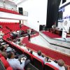 Auditorium with red seats and a panel on stage; audience watching a presentation on stage and screen.