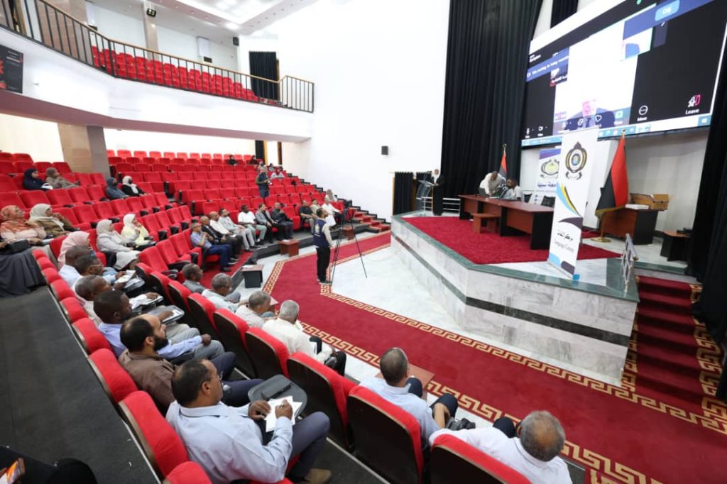 2BDD3A91-1FF7-4414-859E-D23C60C268E3 - جامعة سبها | Sebha University Auditorium with red seats and a panel on stage; audience watching a presentation on stage and screen.