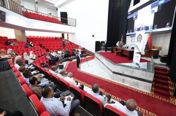 Auditorium with red seats and a panel on stage; audience watching a presentation on stage and screen.