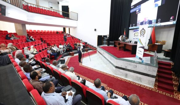 Auditorium with red seats and a panel on stage; audience watching a presentation on stage and screen.