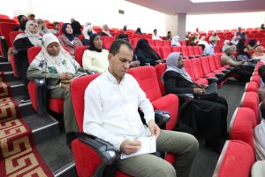 2C1791DE-A217-4EE4-8FB3-E945D7AFBC5A - جامعة سبها | Sebha University Audience in a lecture hall; man in a white shirt writes in a notebook while others sit in red seats behind him, many wearing hijabs.